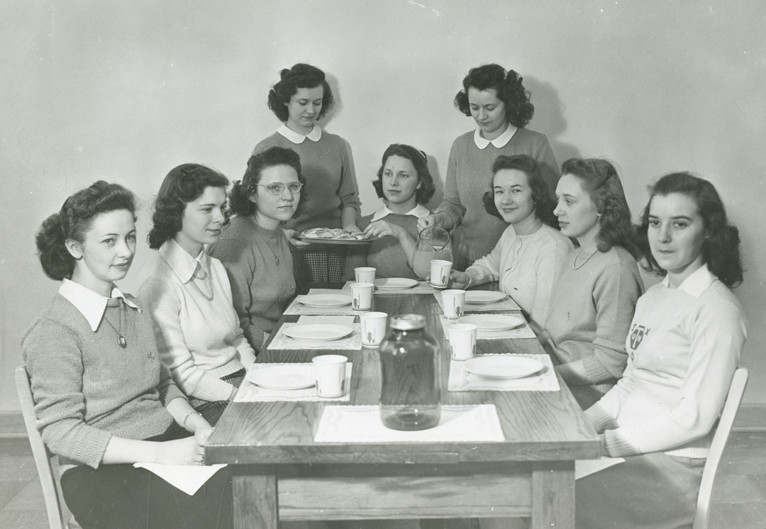 A black and white photograph of women sitting around a wooden table.