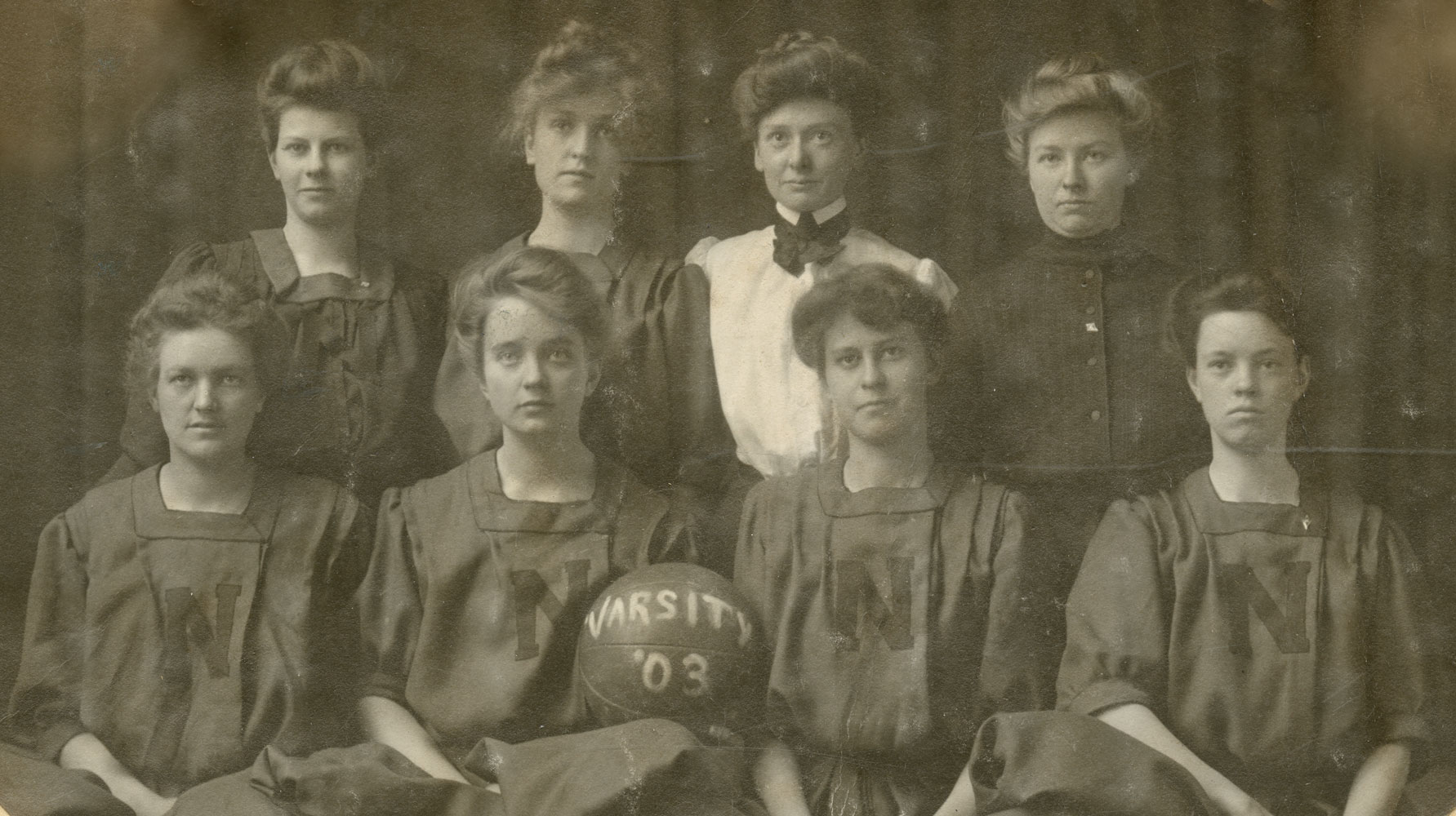 A black and white group portrait of women in dresses holding a basketball