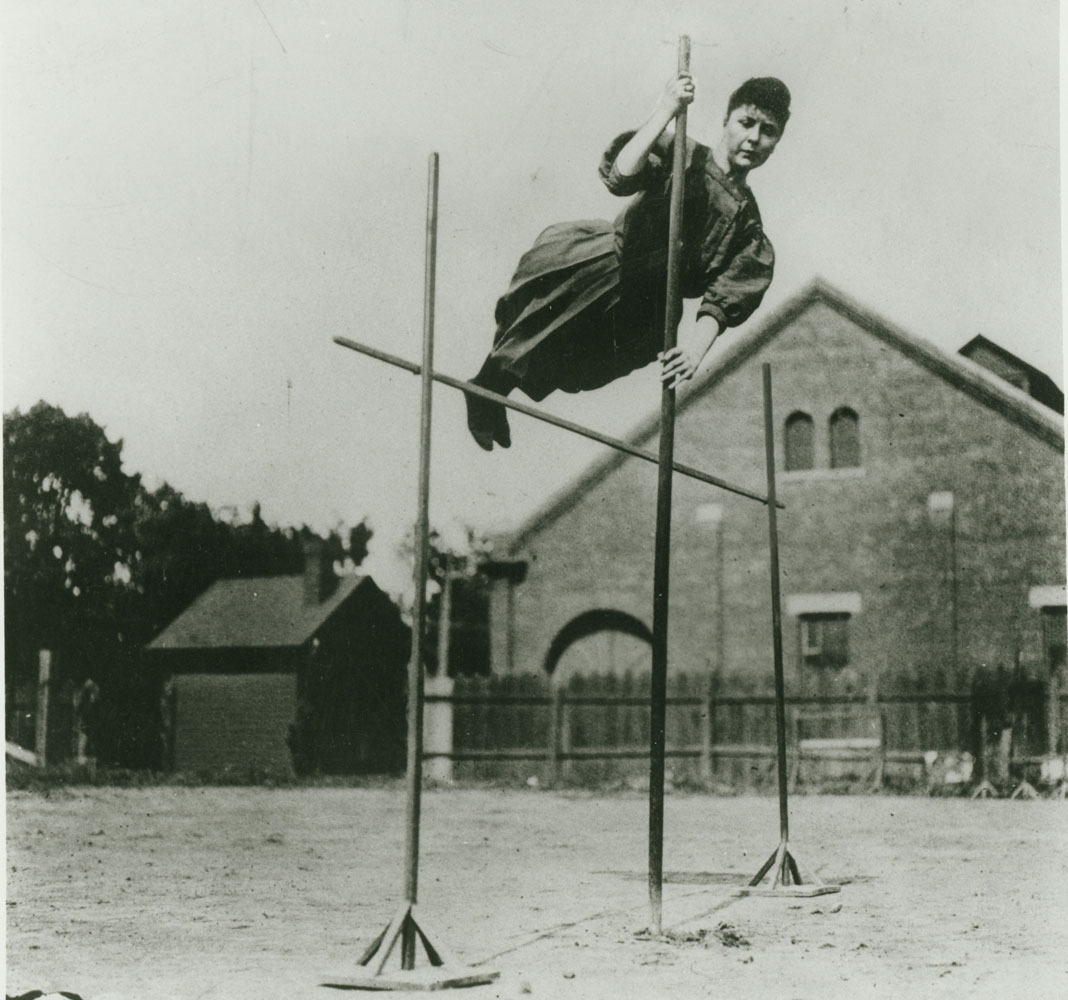 A black and white image of a woman pole vaulting in a dress