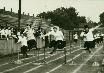 A black and white image of women in skirts jumping over hurdles